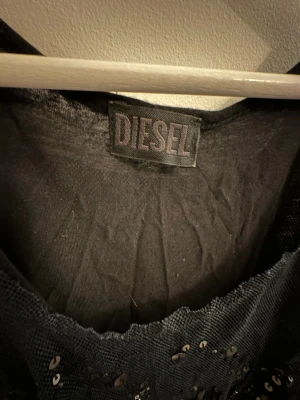 Svart paljettklänning från Diesel - Snygg svart ärmlös klänning från Diesel med transparent mesh och glittriga paljettdetaljer framtill. Kort modell med rund halsringning och loose fit, perfekt för dig som vill sticka ut. Materialet känns lätt och luftigt.