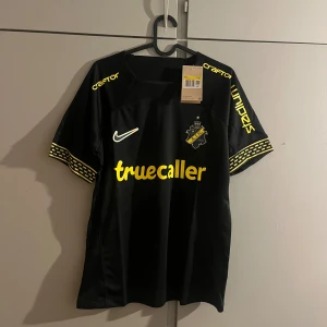 AIK svart matchtröja Nike 2024 - AIK:s officiella matchtröja från Nike, svart med gula detaljer och tryck. Kortärmad med gul text på bröstet och ärmarna, samt klubbmärke och sponsorer. Tillverkad i lätt Dri-FIT-material som andas. Snyggt mönster på ärmsluten och modern passform.