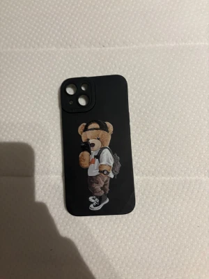 iPhone Skal med Nallemotiv iPhone 15 - Snyggt svart iPhone-skal med ett trendigt nallemotiv. Skalet har öppningar för tre kameror och blixt, passar nyare iPhone-modeller. Skalet ser ut att vara i gott skick utan synligt slitage, perfekt för dig som vill skydda din mobil och ge den en personlig touch.