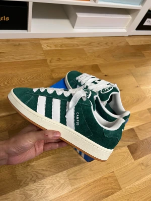 Adidas Campus gröna sneakers mocka - Adidas Campus sneakers i mörkgrön mocka med klassiska vita ränder och vit logga på hälen. Skorna har vit snörning, rund tå och platt sula i gummi med ljusbrun undersida. Stilren retrodesign som passar perfekt till streetwear och casual outfits.