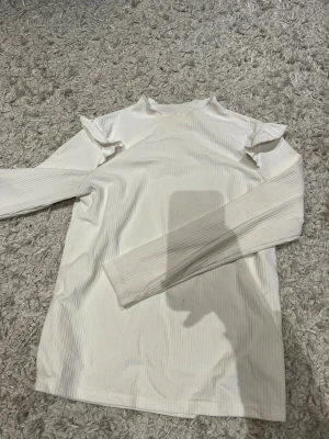 Vit ribbad långärmad tröja Shein - Säljer en vit långärmad tröja från Shein i storlek 11-12 år. Tröjan har ett diskret ribbat mönster och volangdetaljer vid axlarna som ger en söt touch. Passar dig som gillar stilrena och enkla plagg med lite extra detalj.