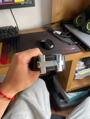 Svart skärp med silverfärgat H-spänne - Snyggt svart bälte med grov textur och vita sömmar längs kanterna. Spännet är stort och i blank silverfärg, format som ett H. Bältet är tillverkat i skinn och har en stilren, modern look som passar till jeans eller kostymbyxor.