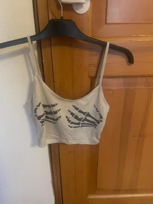 Beige croppad linne med skelettprint - Säljer ett beige croppat linne med tunna axelband och coolt svart skelettprint på framsidan. Perfekt för dig som gillar edgy och unik stil. Materialet känns mjukt och lätt, perfekt för varma dagar eller festivalhäng.