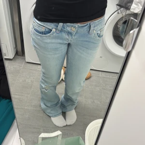 Ljusblå bootcut jeans från G-Rise - Snygga ljusblå jeans från G-Rise med klassisk femficksdesign och bootcut-ben. Jeansen har låg midja, slitning vid knät och kontrastsömmar med detaljerade fickor bak. Perfekt för dig som gillar en avslappnad och trendig look. Storlek S R