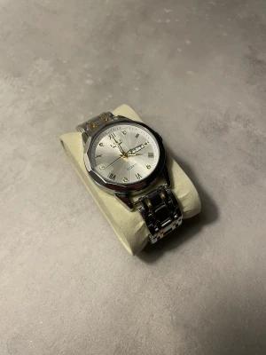 Silverfärgad klocka från Lige - Stilren klocka från Lige med rund urtavla i silver och romerska siffror. Armbandet är i metall med både silver- och guldfärgade länkar. Urtavlan har datumvisning och guldfärgade visare. Perfekt accessoar för dig som gillar klassisk design. Ops: Skaka lite på klockan om den stannat!!