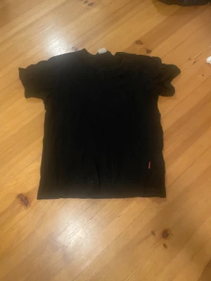 Svart t-shirt från Newbody, storlek L - En klassisk svart t-shirt från Newbody i storlek L. Modellen är enkel med rund hals och korta ärmar, perfekt för en clean och stilren look. Materialet är mjukt och skönt, troligtvis bomull. Diskret röd etikett på sidan ger en liten detalj.