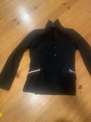 Svart ridkavaj Kingston 160 - Snygg svart ridkavaj från Kingston med broderad logga på fickan och coola röd-vit-svarta detaljer. Kavajen har klassisk krage, knappar framtill och långärmad passform. Perfekt för dig som vill ha stil på tävling eller träning.