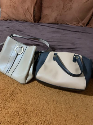 Beige och svart handväska + axelväska - Två snygga väskor säljes! Ena är en beige handväska med svarta detaljer, guldfärgade metalldelar och en vit tag. Den andra är en ljusbeige axelväska med rund guldfärgad ring och dekorativa remmar framtill. Båda har stilren design och är gjorda i skinnimitation.