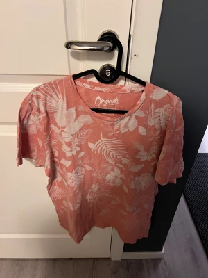 Rosa blommig t-shirt Jack & Jones - Säljer en rosa t-shirt från Jack & Jones Originals med vitt blommigt mönster. T-shirten har rund hals och korta ärmar, perfekt för sommarens chill dagar. Materialet är mjuk bomull som känns skönt mot huden.