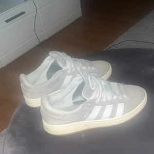 Grå Adidas Campus sneakers i mocka - Snygga Adidas Campus sneakers i ljusgrå mocka med klassiska vita ränder på sidorna och vit sula. Skorna har rund tå, platt sula och snörning. Perfekta för dig som gillar en clean och stilren look med retrovibbar.