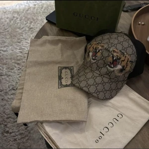 Gucci keps Tiger - Snygg keps från Gucci med klassiskt GG-mönster i beige och brunt, prydd med två coola tigrar framtill. Svarta mesh-paneler på sidorna och justerbar rem bak med röd-gröna detaljer. Perfekt för dig som vill sticka ut med lyxig streetstyle.