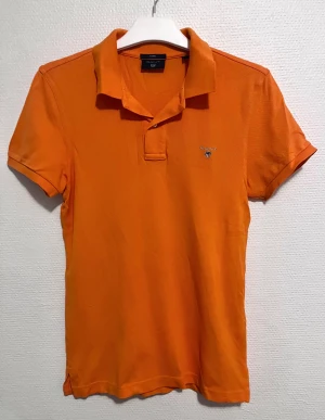 Orange pikétröja från GANT XS FITTED - Snygg orange pikétröja från GANT med klassisk krage och korta ärmar. Tröjan har broderad logga på bröstet och är tillverkad i 100% bomull med en skön, lite grov struktur. Perfekt för dig som gillar färg och vill ha en stilren look.