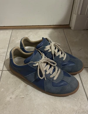 Blå sneakers Maison Margiela - Snygga blå Maison Margiela sneakers i mocka och skinn, beige snörning och klassisk gummisula. Skorna har en retroinspirerad design med rund tå och diskreta detaljer. Perfekta för dig som gillar stilrena och tidlösa sneakers.