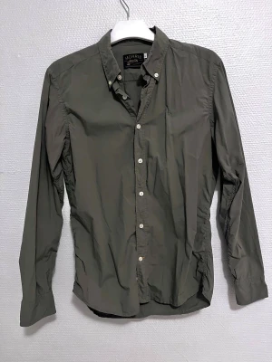 Olivgrön skjorta från Morris, S - Olivgrön skjorta från Morris med klassisk krage och vita knappar framtill. Långärmad modell i 100% mjuk bomull, med broderad logga på bröstet. Snygg och stilren passform som funkar till många olika looks. Perfekt för dig som gillar enkel och clean stil.