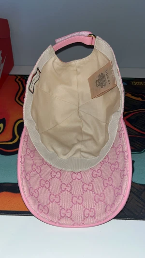 Rosa Gucci keps med GG-mönster för baseball - Snygg rosa keps från Gucci med klassiskt GG-monogram över hela kepsen. Justerbar rem bak i rosa skinn och detaljer i guld. Materialet är bomull med inslag av skinn, och kepsen har en rundad form med böjd skärm. Perfekt för dig som vill sticka ut med lyxig streetstyle.