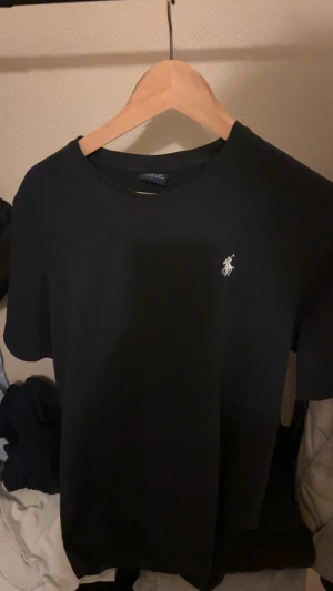 Svart t-shirt från Polo Ralph Lauren - Svart t-shirt från Polo Ralph Lauren med klassisk rund hals och vit broderad logga på bröstet. Enkel och stilren design, perfekt för dig som gillar cleana plagg med lite lyxig känsla. Mjuk och skön bomull som sitter snyggt på kroppen.