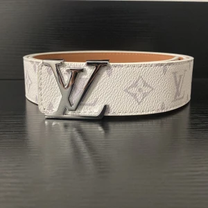 Vit skärp i skinn - Säljer ett snyggt vitt bälte i skinn med klassiskt monogrammönster och en stor silverfärgad logga Perfekt för dig som vill sticka ut med en lyxig accessoar.