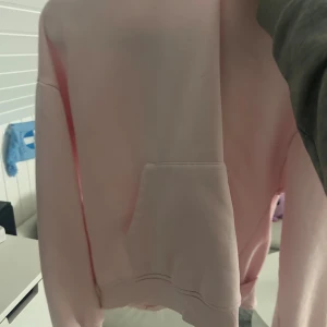 Ljusrosa hoodie med ficka - Säljer en ljusrosa hoodie med stor magficka och ribbade muddar. Tröjan har en klassisk, avslappnad passform och är perfekt för chill dagar. Mjuk och skön, passar dig som gillar enkel och stilren streetwear.