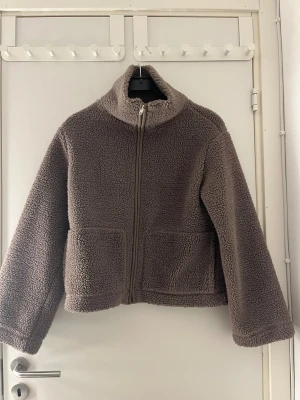 Brun teddyjacka med dragkedja - Mysig brun teddyjacka med hög krage och två stora fickor framtill. Jackan har en rak och loose passform, långärmade ärmar och stängs med dragkedja. Perfekt för att hålla sig varm och se chill ut under höst och vinter.