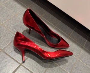Röda pumps från Zara Basic Collection - Snygga röda pumps med metallicfinish från Zara Basic Collection. Skorna har spetsig tå, låg klack och en elegant, glansig yta som verkligen sticker ut. Insidan är svart och de har en klassisk siluett som ger en cool vibe till din outfit. Syns som nya. 