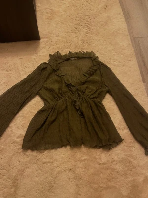 Olivgrön blus med volangdetaljer - Säljer en olivgrön/khaki blus med lång ärm och v-ringning. Blusen har volangdetaljer längs ringningen och framtill, supersnygg!💚