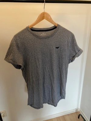 Grå t-shirt från Hollister -storlek Medium - Snygg grå t-shirt från Hollister med klassisk rund hals och liten svart logga på bröstet. T-shirten är i mjuk bomull och har korta ärmar med uppvik. Perfekt för dig som gillar enkel och stilren look.