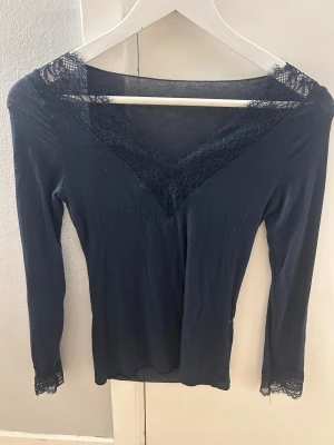 Mörkblå långärmad topp med spets - Säljer en mörkblå långärmad topp med v-ringad hals och fina spetsdetaljer från intimissmi. Toppens material är mjukt och följsamt, perfekt för dig som gillar en elegant och stilren look. Passar dig som vill ha något extra till outfiten.