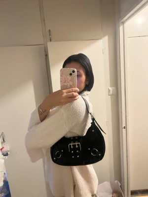 Svart axelväska med stora metalldetaljer - Cool svart axelväska i syntet med chunky bälte och silvriga nitar. Väskan har flera ringar och detaljer i metall, samt dragkedja upptill. Perfekt storlek för det viktigaste och ger en edgy vibe till din outfit.