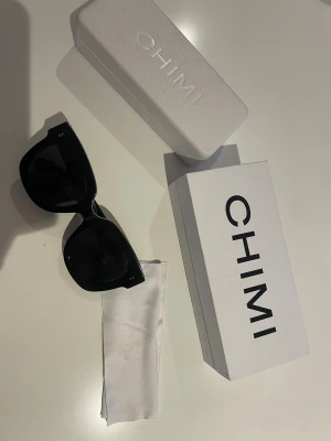 Svarta CHIMI solglasögon med box - Snygga svarta solglasögon från CHIMI med breda, fyrkantiga bågar i blankt syntetmaterial. Kommer med original vit box och putsduk. Perfekt accessoar för att höja din stil och skydda ögonen mot solen. Model 08, Solglasögona är varsamt använda, jag har ej hittat några defekter, men kan såklart förekomma något litet som jag missat