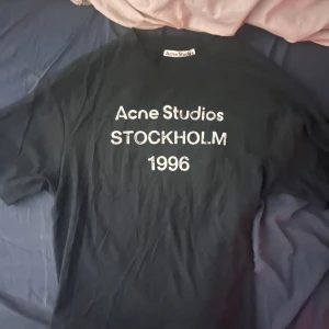 Svart Acne Studios t-shirt Stockholm - Svart t-shirt från Acne Studios med vit text 'Acne Studios STOCKHOLM 1996' på bröstet. Klassisk rund hals och korta ärmar. Materialet är mjuk bomull och passformen är normal. Perfekt för dig som gillar stilrena och ikoniska plagg. Passar runt 170-180 ungefär