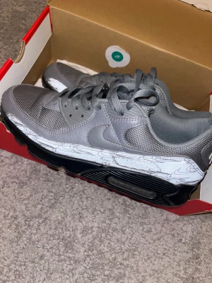 Grå Nike Air Max 90 sneakers - Säljer ett par grå Nike Air Max 90 sneakers med svart sula och snygga detaljer på sidan. Skorna har mesh och skinnpaneler, snörning och en platt sula med synlig Air-enhet. Perfekt för dig som gillar stilrena och sportiga sneakers med unik look.