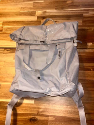Ljusgrå ryggsäck från Nike - Snygg ljusgrå ryggsäck från Nike med stjärnmönster och 'JUST DO IT'-tag på framsidan. Väskan har både handtag och justerbara axelremmar, samt en stor ficka framtill. Tillverkad i slitstark polyester och har en stilren look med diskreta Nike-loggor.