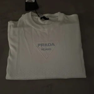 Vit t-shirt från Prada med logga - Vit t-shirt från Prada med klassisk rund hals och korta ärmar. Framsidan har en snygg, broderad Prada Milano-logga i triangelform. Materialet känns mjukt och exklusivt, perfekt för dig som gillar stilrena designer och lyxiga märken. Äkta med prislapp. Aldrig använd 