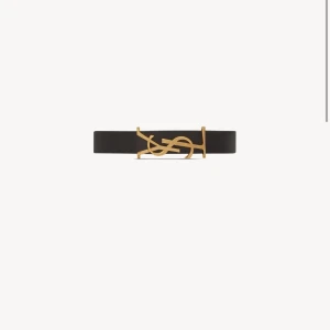 Svart YSL bälte med guldspänne - Supersnyggt svart bälte från Saint Laurent med ikoniskt YSL-spänne i guld. Bältet är tillverkat i slätt skinn och har en stilren, klassisk form som lyfter vilken outfit som helst. Perfekt accessoar för dig som gillar lyxiga detaljer och designer. Använt några gånger och nypris på hemsidan är cirka 3000