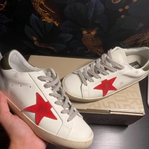 Golden Goose Superstar sneakers vit/röd - Golden Goose Superstar sneakers i vitt skinn med en röd stjärna på sidan och grå snörning. Bakdelen har en olivgrön detalj. Klassisk rund tå och platt sula. Snygg italiensk design med coola street vibes, perfekt för dig som gillar att sticka ut.