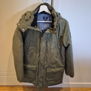 Grön parkajacka från GANT med huva - Snygg grön parkajacka från GANT med stor huva och praktiska fickor framtill. Jackan har dragkedja och tryckknappar, samt justerbar huva med snörning. Perfekt för kyliga dagar och har en klassisk, rak passform.