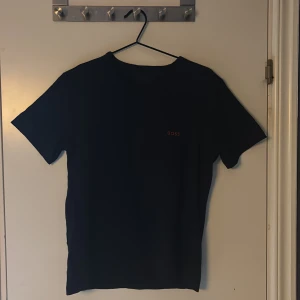 Svart BOSS t-shirt med röd logga - Svart t-shirt från BOSS i storlek M med stilren röd broderad logga på bröstet. Klassisk rund hals och korta ärmar. Materialet är mjuk bomull som känns skönt mot huden. Perfekt för dig som gillar minimalistisk design och snygga detaljer.