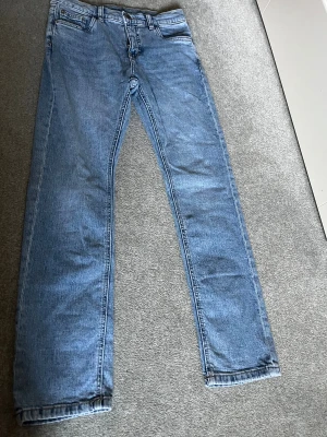 Blå jeans från Kappahl, storlek 152 - Snygga ljusblå jeans från Kappahl i klassisk femficksmodell. Jeansen har rak passform och normal midja, med dragkedja och knapp framtill. Perfekta för dig som gillar enkel och stilren denim-look.
