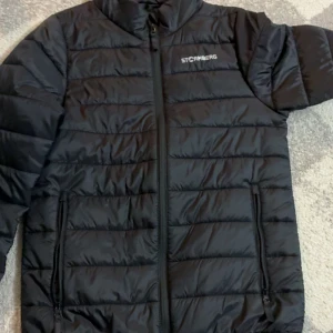 Svart pufferjacka från Stormberg - Svart pufferjacka från Stormberg med horisontella quiltade sömmar och dragkedja framtill. Jackan har två sidofickor med dragkedja och en hög krage. Materialet är syntet och jackan är perfekt för kyliga dagar.