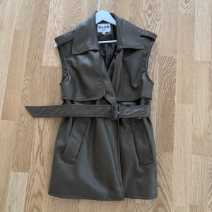 Mörkbrun skinnväst från NA-KD - Snygg mörkbrun skinnväst från NA-KD med bred krage och bälte i midjan. Västen har två framfickor och en stilren, rak passform. 
