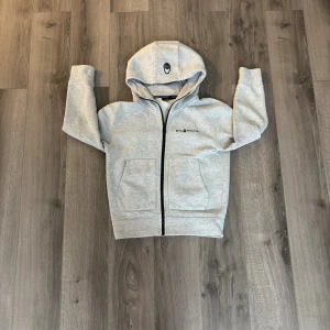 Grå hoodie från Sail Racing storlek 150 barn - Snygg ljusgrå hoodie från Sail Racing med svart logga på bröstet och huvan. Tröjan har dragkedja framtill, två stora fickor och en skön huva. Perfekt för chill dagar och har en clean look som funkar till jeans eller joggers. Storlek 150 barn