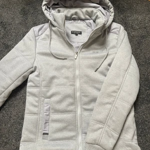 Ljusgrå jacka från Koroshi - Snygg ljusgrå jacka från Koroshi med huva och dragsko. Jackan har quiltad design, dragkedja framtill och två fickor med detaljer i blankare tyg. Diskret logga på bröstet och mjukt syntetmaterial som håller värmen. Perfekt för kyliga dagar. Hör av er vid frågor eller funderingar!🤩