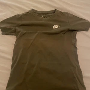 Olivgrön Nike t-shirt med logga - Snygg olivgrön t-shirt från Nike med klassisk vit logga på bröstet. Modellen har rund hals och korta ärmar, perfekt för en chill och sportig look. Tillverkad i mjuk bomull som känns skön mot huden.