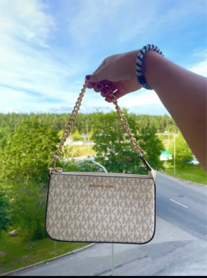 Michael Kors beige handväska med kedja - Snygg beige handväska från Michael Kors med MK-logga över hela väskan. Väskan har guldfärgad kedja som axelrem och dragkedja upptill. Insidan har fack för kort och småsaker. Perfekt storlek för det viktigaste och stilren design.