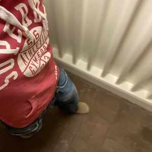 Superdry Hoodie - Säljer en röd hoodie från Superdry med stort vitt tryck framtill och på ärmen, sälj i befintlig skick. Tröjan har huva med vita snören och en klassisk känguruficka. Perfekt för dig som gillar sportig och avslappnad stil. Om du undrar något är det bara att skriva🙌✅