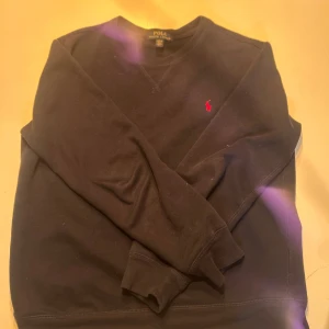 Svart sweatshirt från Polo Ralph Lauren - Svart sweatshirt från Polo Ralph Lauren med klassisk röd broderad logga på bröstet. Tröjan har rund hals, långa ärmar och är gjord i mjuk bomull med lite polyester för extra komfort. Perfekt för chill dagar och enkel att matcha med jeans eller joggers. PRIS KAN DISKUTERAS.
