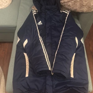 Blå Adidas pufferjacka med huva - Säljer en mörkblå pufferjacka från Adidas med vit logga på bröstet och klassiska vita ränder längs axlarna. Jackan har huva, dragkedja och vita detaljer på sidorna. Perfekt för kalla dagar och sportig stil.