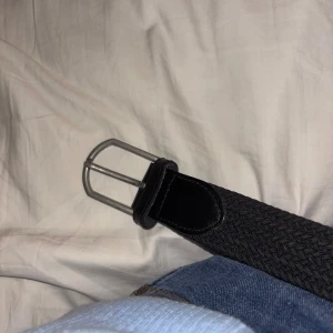 Svart flätat bälte med silverfärgat spänne - Säljer ett svart flätat bälte med silverfärgat metallspänne. Bältet har detaljer i svart skinn vid ändarna och en snygg, enkel design som passar till jeans eller andra byxor. Perfekt accessoar för att lyfta din outfit.