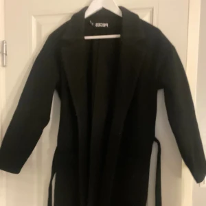 Svart kappa med bälte från Pieces - Svart lång kappa från Pieces med klassisk krage och bälte i midjan. Stilren och enkel design med raka linjer och långa ärmar i storlek S. kappan är oanvänd endast provad fåtal gång. Pris går att diskutera vid snabb affär och har möjlighet att skicka samma dag😍