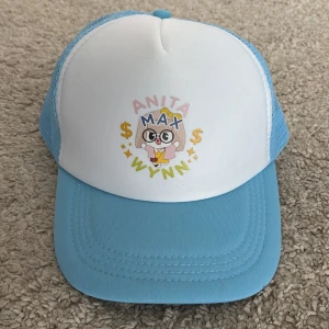 Anita Max Wynn keps  - Säljer en blå och vit keps med mesh på sidorna och justerbar snapback. Framtill finns ett färgglatt tryck med texten 'ANITA MAX WYNN' och en söt tecknad figur med glasögon, rosa jacka och gul klänning. Perfekt för dig som gillar roliga och unika accessoarer.